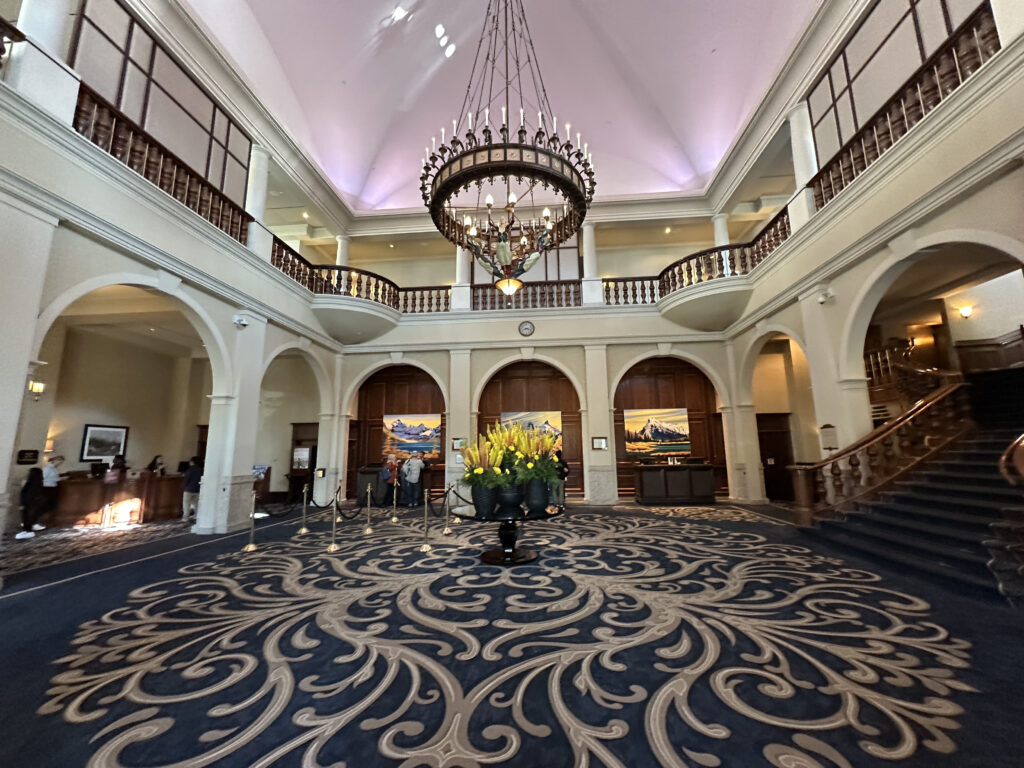 The image shows a grand hotel lobby with a large, ornate chandelier hanging from a high ceiling. The space features a patterned carpet and a central floral arrangement. There are two levels with a balcony and wooden railings. On the left, there is a reception desk with people standing nearby. On the right, a staircase leads to the upper level. The walls are adorned with paintings, and the overall atmosphere is elegant and spacious.