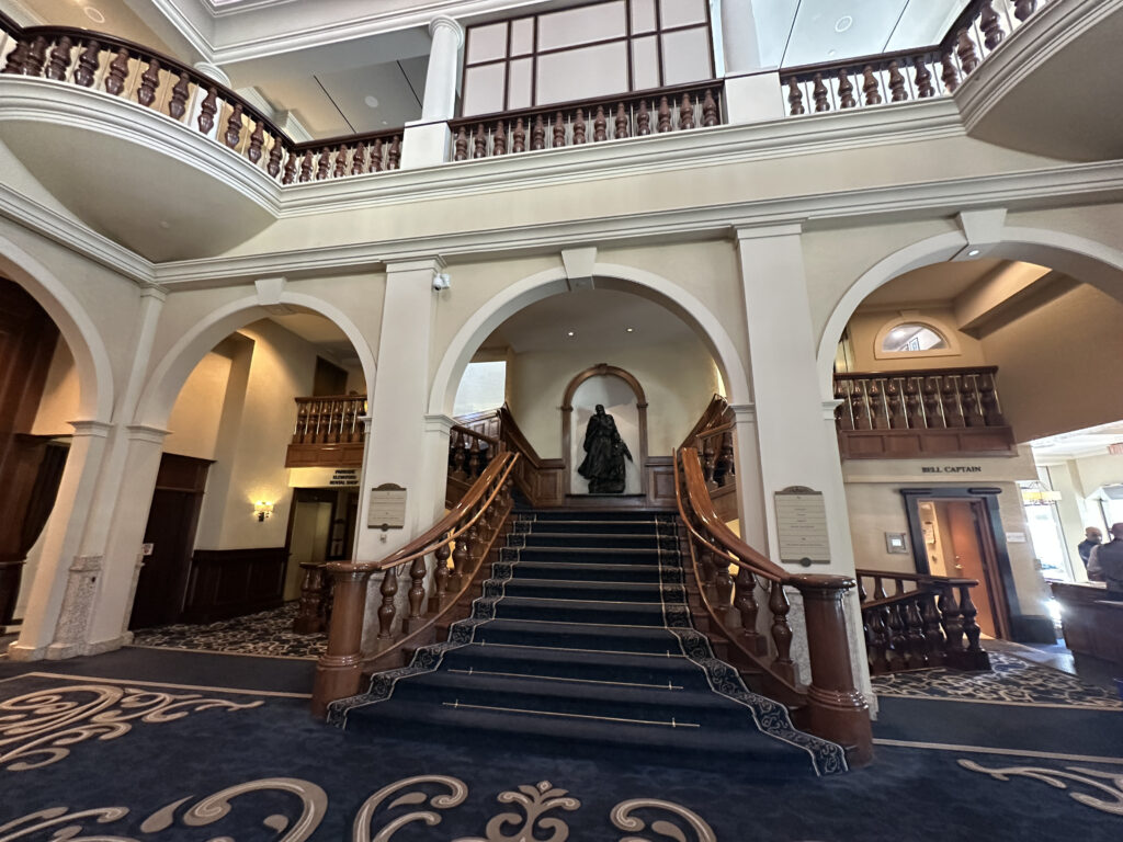 The image shows an elegant interior of a building with a grand staircase. The staircase is carpeted in dark blue with ornate wooden railings. Above the staircase, there is a statue in an alcove. The surrounding architecture features arches and decorative moldings. There are two levels visible, with a balcony on the upper level. The floor is covered with a patterned carpet, and there are signs indicating directions, including one for a "Bell Captain."
