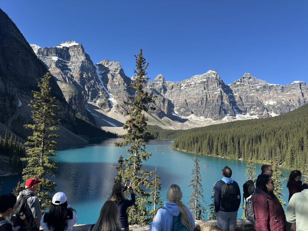 A group of people is standing on a viewpoint overlooking a stunning turquoise lake surrounded by dense evergreen forests and towering snow-capped mountains under a clear blue sky. Some people are taking photos of the scenic landscape.