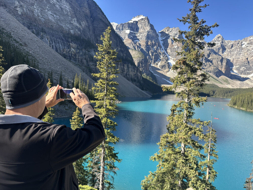 A person wearing a black jacket and beanie is taking a photo with a smartphone of a stunning landscape. The scene features a turquoise lake surrounded by tall evergreen trees and rugged mountains with patches of snow under a clear blue sky.