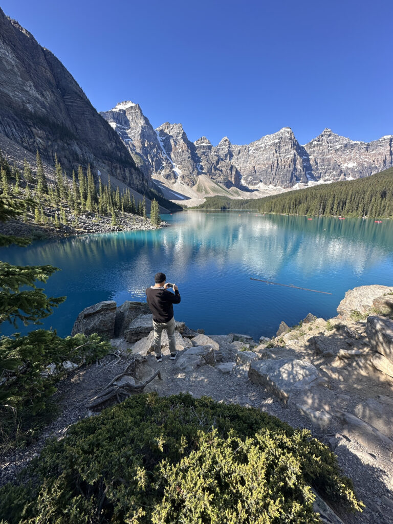 A person is standing on a rocky ledge, taking a photo of a stunning mountain landscape. The scene includes a clear blue lake surrounded by tall, rugged mountains with patches of snow. Pine trees line the shore, and the sky is clear and bright blue.