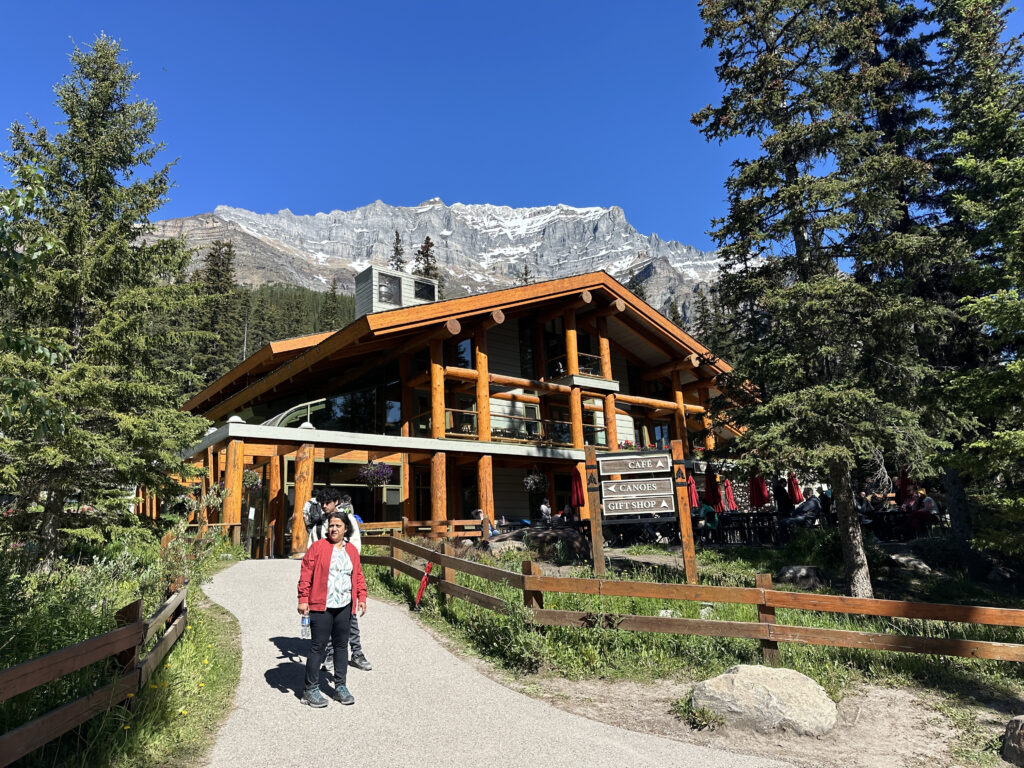 A wooden lodge with large windows is surrounded by tall pine trees and set against a backdrop of a clear blue sky and a snow-capped mountain. A person in a red jacket is walking on a path leading to the lodge. There is a wooden signpost indicating directions to a cafe, canoes, and a gift shop. The area is lush with greenery and the scene is bright and sunny.