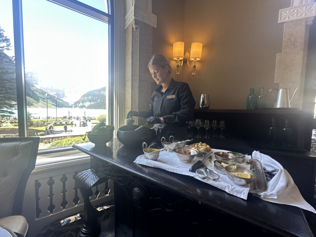 A person is preparing food at a table inside a restaurant. The table is set with various dishes and utensils. There is a large window with a view of a scenic landscape featuring mountains, a lake, and people walking outside. The interior is warmly lit with wall sconces.