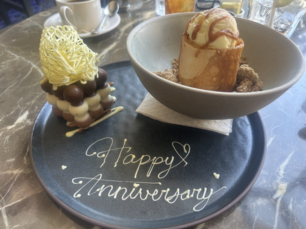 A dessert plate with "Happy Anniversary" written in white chocolate. On the left, there's a stack of chocolate spheres topped with a white chocolate lattice. On the right, a bowl contains a rolled pastry with ice cream and caramel sauce, surrounded by crumbled toppings. A cup and saucer are in the background.
