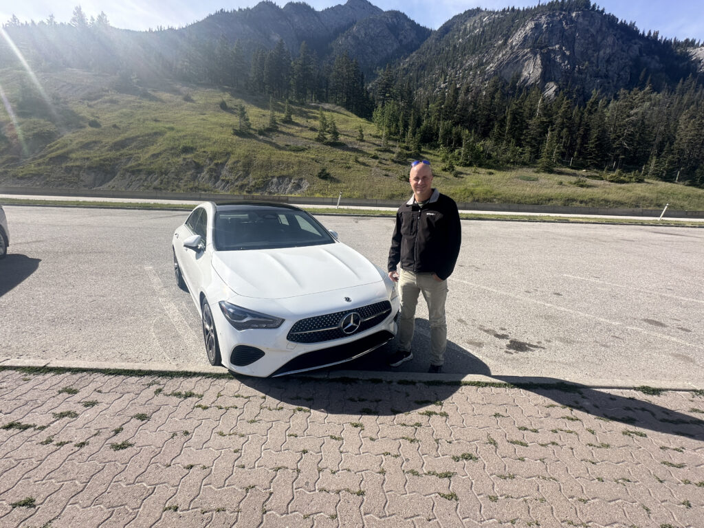 A person is standing next to a white Mercedes-Benz car in a parking area. The background features a scenic view of mountains and trees under a clear sky.