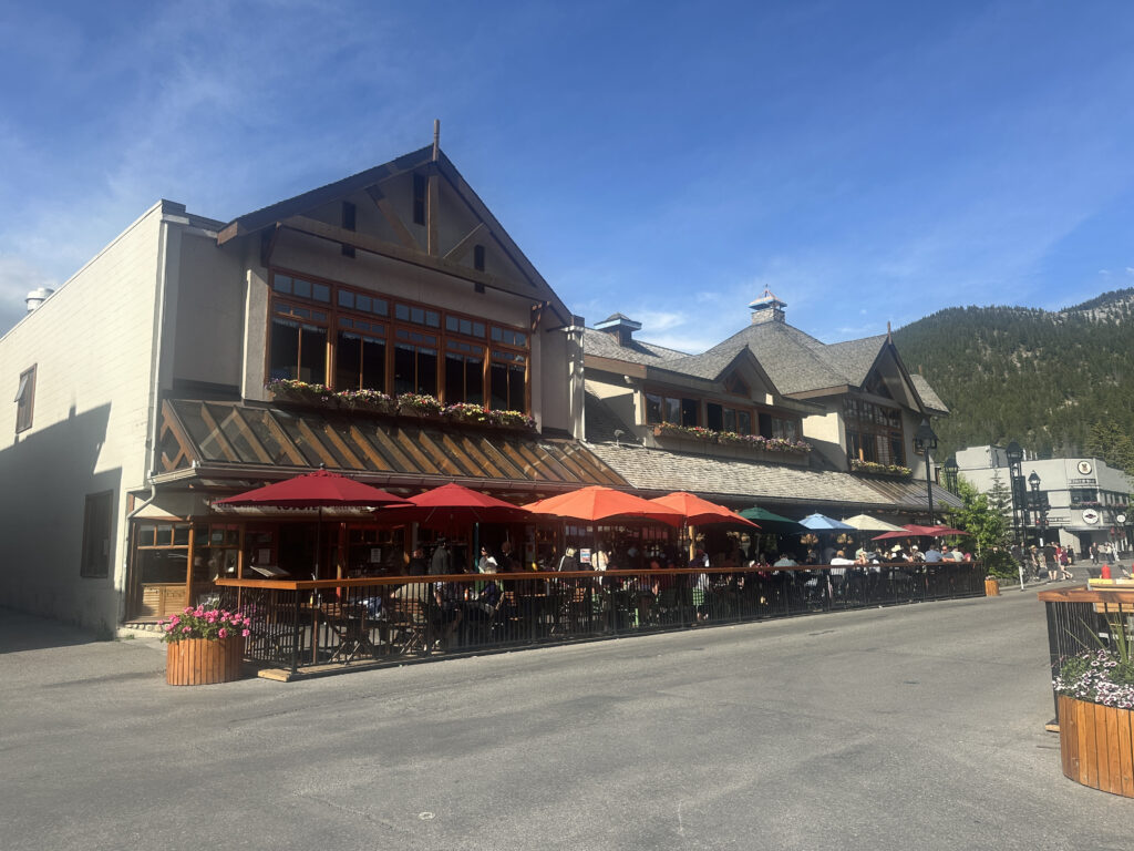The image shows a two-story building with a rustic design, featuring large windows and flower boxes. There is an outdoor seating area with tables and colorful umbrellas in red, orange, green, and blue. People are sitting and enjoying the sunny weather. The building is set against a backdrop of a clear blue sky and a forested mountain. The street in front is lined with flower planters.