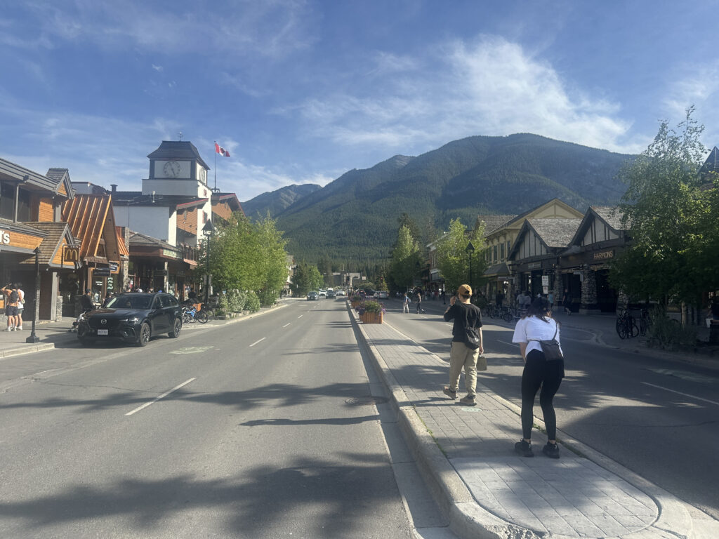 A street scene in a small town with a mountain backdrop. The street is lined with shops and buildings, including a clock tower and a McDonald's. A Canadian flag is visible on the clock tower. There are people walking on the sidewalk and a few cars parked along the street. The sky is clear and blue.