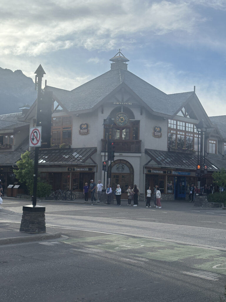 A two-story building with a peaked roof and a clock tower is shown. The building houses a Starbucks and other shops. Several people are standing on the sidewalk in front of the building. There is a no left turn traffic sign and a bicycle rack nearby. The sky is partly cloudy, and mountains are visible in the background.