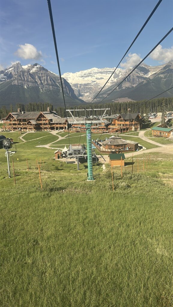 A scenic view from a ski lift overlooking a resort area with wooden lodges and buildings surrounded by green grass. In the background, there are tall, snow-capped mountains under a clear blue sky.