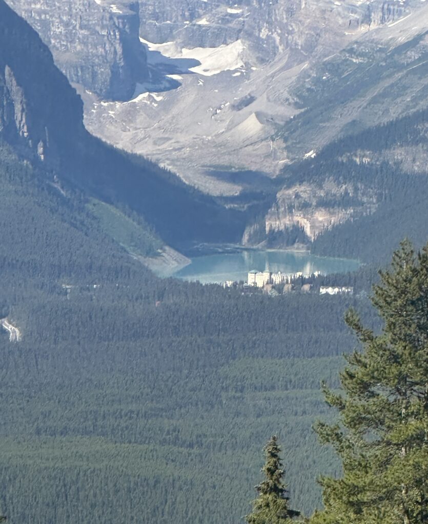A scenic view of a mountainous landscape with a turquoise lake nestled between forested hills. In the foreground, there are dense green trees, and in the distance, a large building is visible near the lake's edge. The background features rugged mountain peaks with patches of snow.
