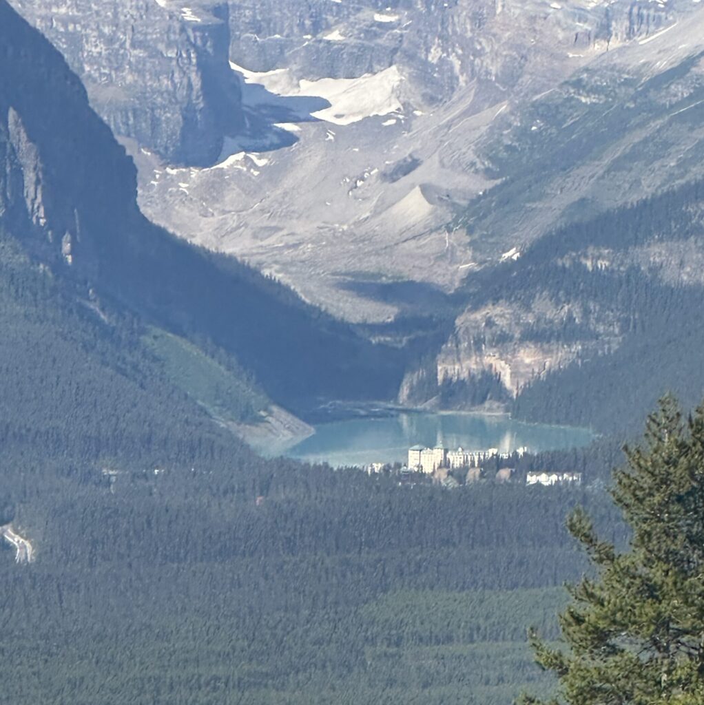 A scenic view of a mountainous landscape with a turquoise lake nestled between forested hills. In the foreground, there are dense green trees, and in the distance, a large building is visible near the lake's edge. The background features rugged mountain peaks with patches of snow.