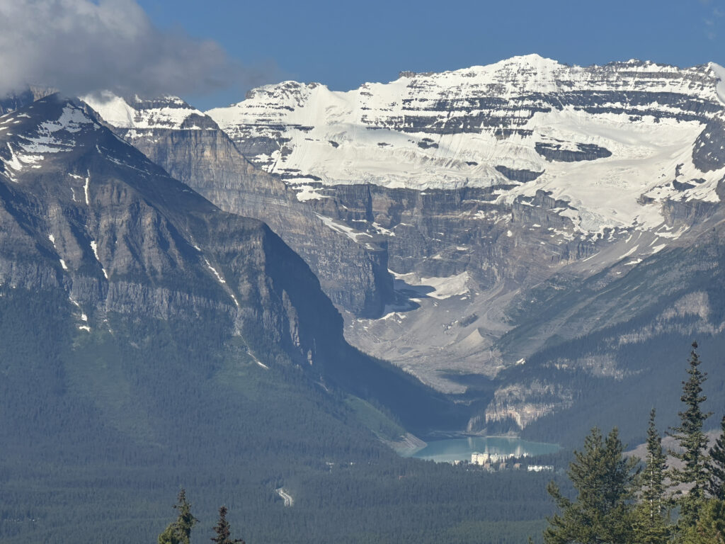 A scenic view of a mountainous landscape featuring snow-capped peaks under a clear blue sky. In the foreground, there are dense green forests, and a turquoise lake is visible at the base of the mountains. The lake is surrounded by trees, and a building can be seen near its shore.
