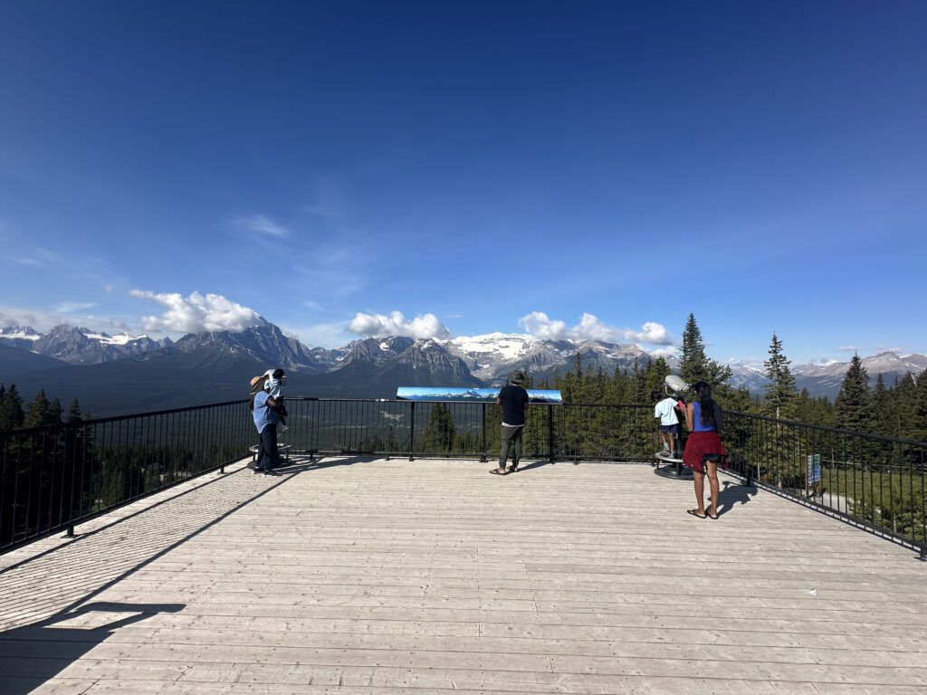 A group of people is standing on a wooden observation deck with a metal railing, overlooking a scenic view of snow-capped mountains and a forested valley. The sky is clear and blue, with a few clouds scattered above the mountains.