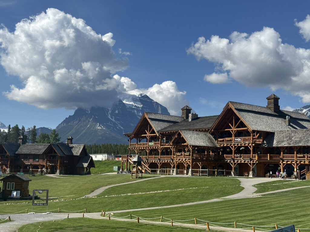 A scenic view of a large wooden lodge with multiple peaked roofs, set against a backdrop of a snow-capped mountain and a blue sky with fluffy clouds. The foreground features a well-maintained lawn with winding pathways.