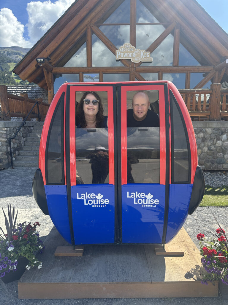 The image shows two people sitting inside a stationary gondola cabin display labeled "Lake Louise Gondola." The gondola is red and blue, with the Lake Louise logo on it. Behind them is a wooden building with a sign that reads "Corona Powder Keg Lounge." There are flower arrangements on either side of the gondola, and the sky is partly cloudy.