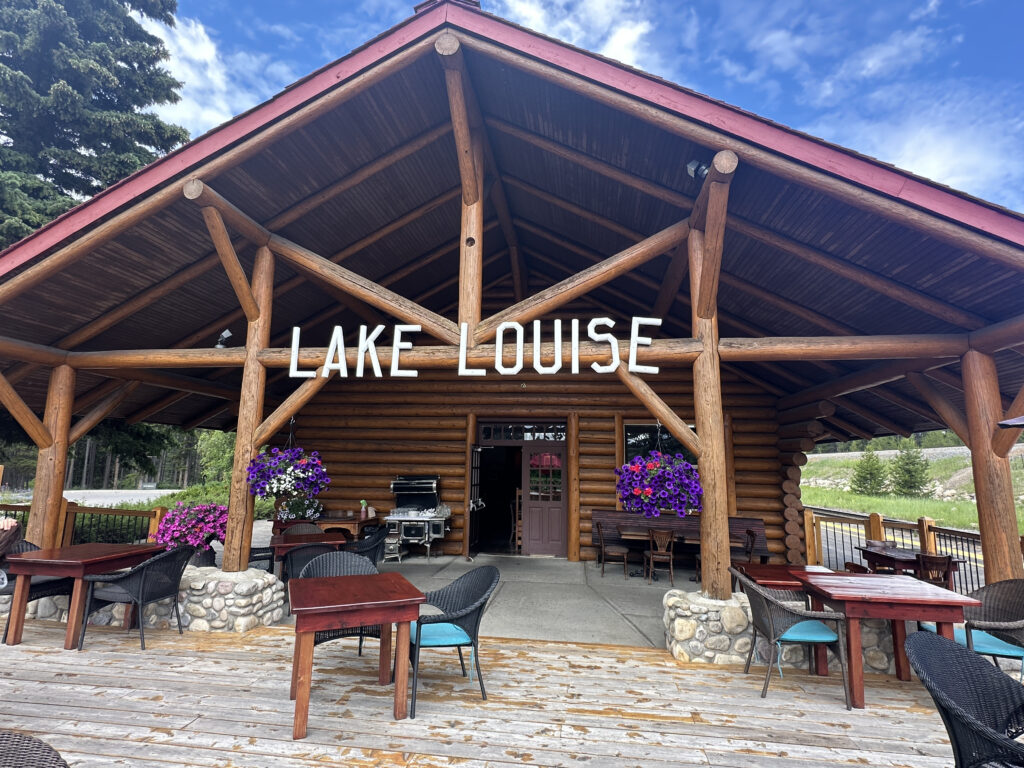 A rustic log cabin with a large wooden sign reading "Lake Louise" is shown. The cabin has a wooden deck with several tables and chairs. Hanging baskets of purple flowers decorate the entrance. The background features trees and a partly cloudy sky.