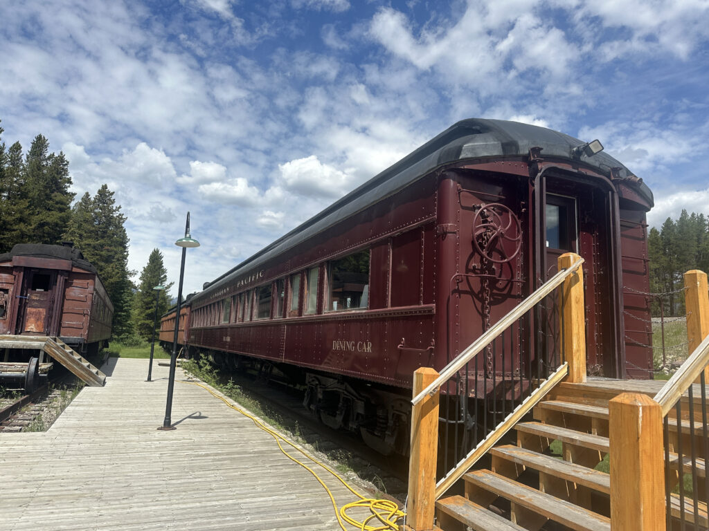 The image shows a vintage red train car labeled "Canadian Pacific" and "Dining Car" on a railway track. There is a wooden platform with stairs leading up to the train car. Another train car is visible in the background. The scene is set outdoors with a clear blue sky and some clouds, surrounded by trees.