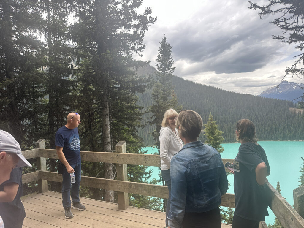 A group of people stands on a wooden viewing platform overlooking a scenic landscape. The background features a turquoise lake surrounded by dense evergreen forests and mountains under a cloudy sky. The people are casually dressed, and one person is holding a water bottle.