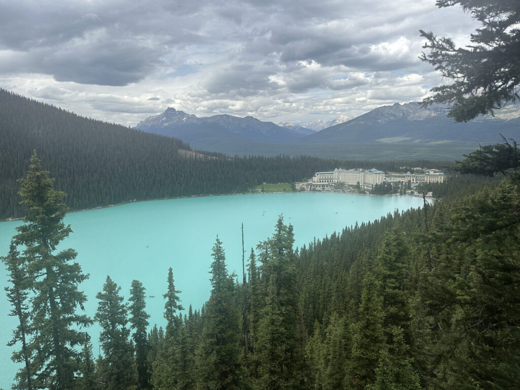 A scenic view of a turquoise lake surrounded by dense evergreen forests and mountains in the background. A large hotel complex is situated on the shore of the lake. The sky is overcast with clouds, adding a dramatic effect to the landscape.