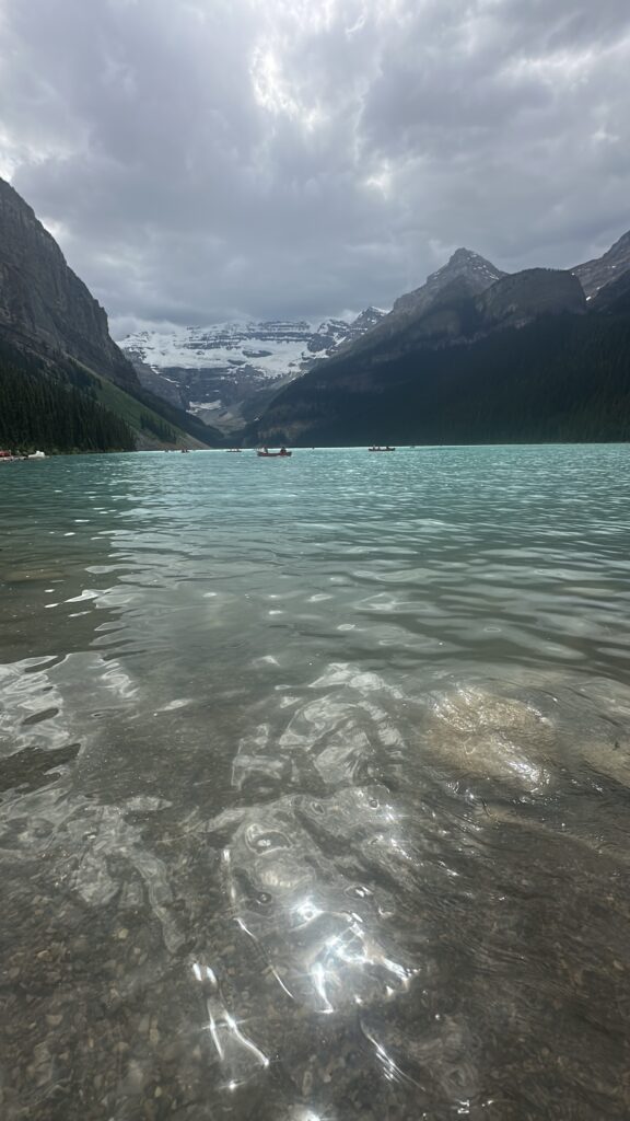 A serene lake with clear turquoise water reflecting the cloudy sky. In the background, there are snow-capped mountains and dense forests lining the shore. Small boats are visible on the lake, adding a sense of scale to the expansive natural landscape.