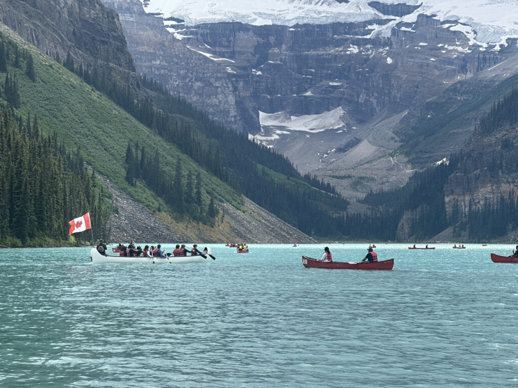 A scenic view of a turquoise lake surrounded by forested mountains and snow-capped peaks. Several canoes are on the water, with people paddling. One canoe prominently displays a Canadian flag. The sky is overcast, adding a serene atmosphere to the landscape.
