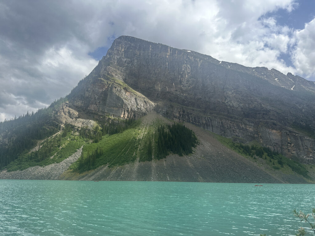 A scenic view of a large mountain with rocky cliffs and patches of greenery, rising above a turquoise lake. The sky is partly cloudy, with sunlight illuminating parts of the mountain. A small boat is visible on the lake.