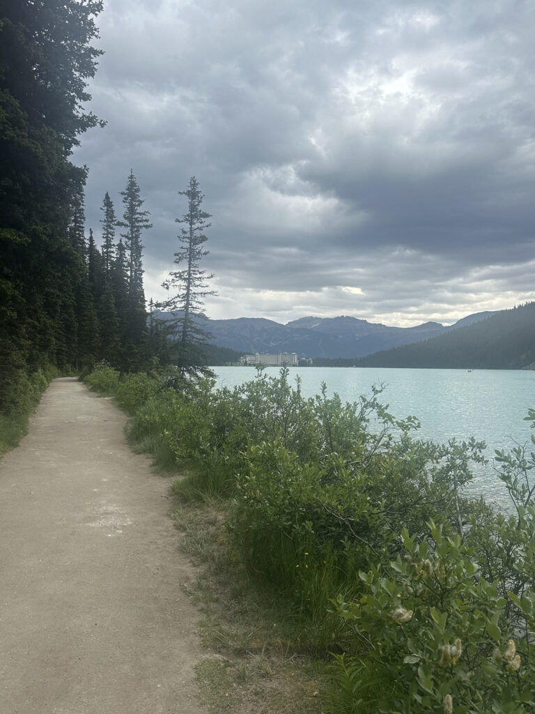 A scenic view of a lakeside path with lush greenery on both sides. The path runs alongside a calm, turquoise lake, with tall evergreen trees lining the left side. In the background, there are mountains under a cloudy sky, creating a serene and picturesque landscape.