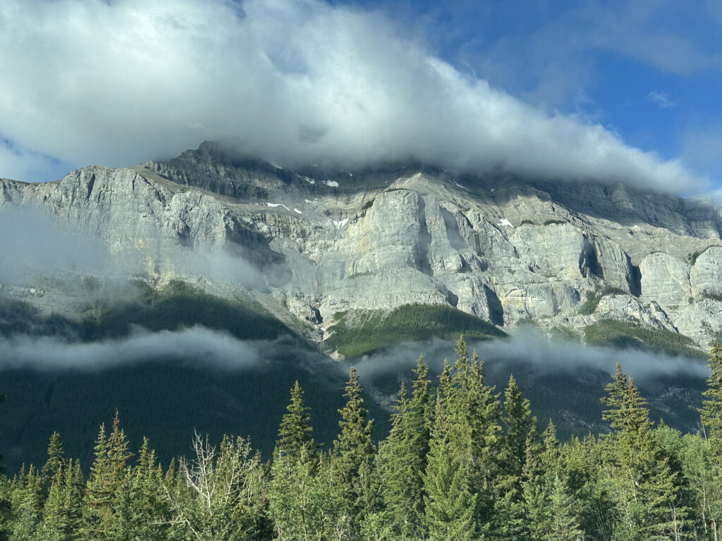 A scenic view of a mountain range partially covered by clouds. The rocky peaks are visible above a dense forest of evergreen trees. The sky is mostly clear with some clouds, creating a dramatic and picturesque landscape.