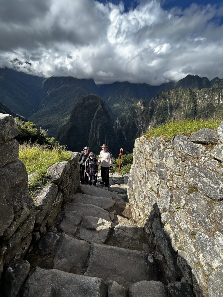 A group of people is walking up a stone staircase surrounded by ancient stone walls. In the background, there are lush green mountains under a cloudy sky. The scene appears to be set in a mountainous, historical location.