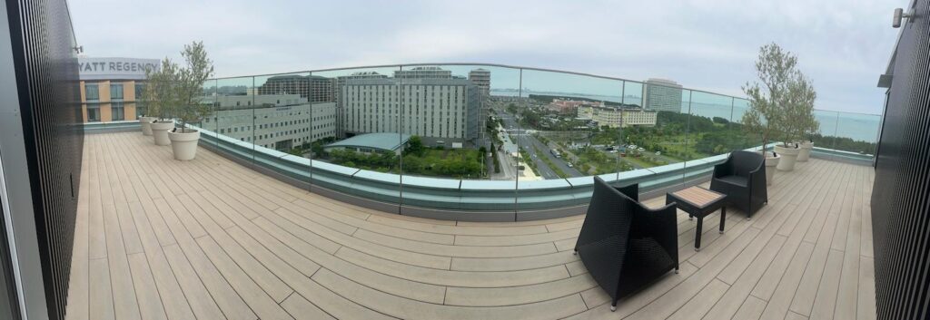 The image shows a rooftop terrace with wooden flooring and glass railings. There are two black wicker chairs and a small table on the right side. Several potted trees line the edge of the terrace. In the background, there is a view of a cityscape with multiple buildings, roads, and greenery, leading to a body of water under a cloudy sky.