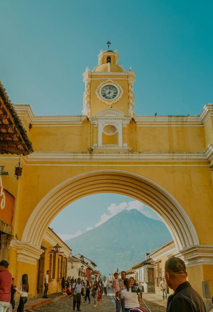 A yellow archway with a clock tower stands prominently in the center of a cobblestone street. People are walking and taking photos beneath the arch. In the background, a large mountain is visible under a clear blue sky.