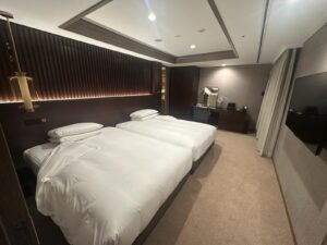 The image shows a modern hotel room with two neatly made twin beds featuring white linens. The room has a dark wood headboard with ambient lighting. There is a desk with a mirror, chair, and some electronic devices on the right side. A flat-screen TV is mounted on the wall opposite the beds. The room has a neutral color scheme with beige carpet and curtains.