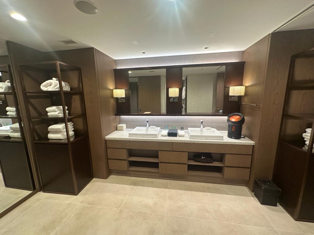 The image shows a modern bathroom with a double sink vanity. The vanity has a large mirror above it, flanked by two wall-mounted lights. On the countertop, there are two sinks with a few toiletries in between. To the left and right of the vanity, there are open shelves stocked with neatly folded white towels. The floor is tiled, and there is a small trash bin on the right side.