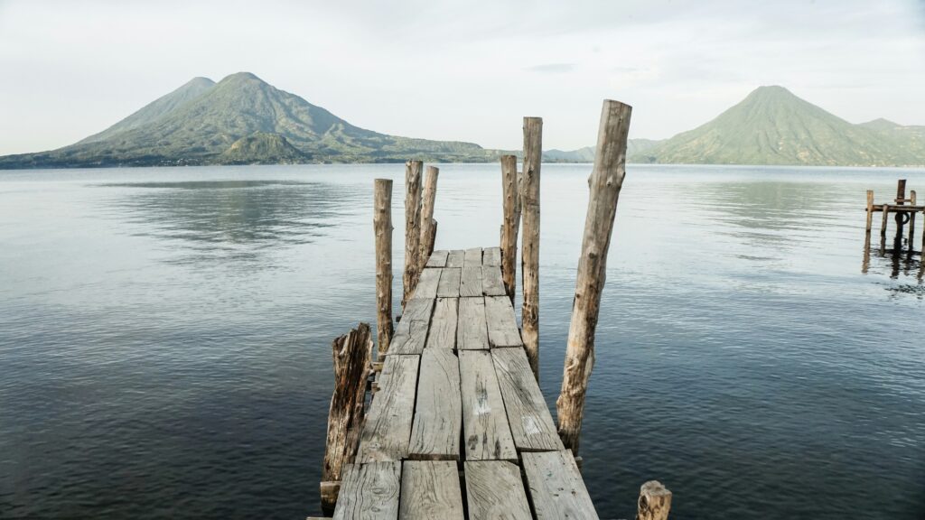 A wooden pier extends over a calm lake, leading towards two large, green mountains in the background. The sky is overcast, and the water reflects the mountains and sky, creating a serene and peaceful scene.