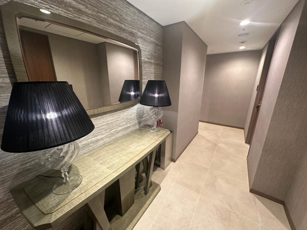 The image shows a modern hallway with a textured wall and a large rectangular mirror. Below the mirror is a console table with a glass lamp featuring a black lampshade. The floor is tiled, and the hallway is well-lit with recessed ceiling lights.