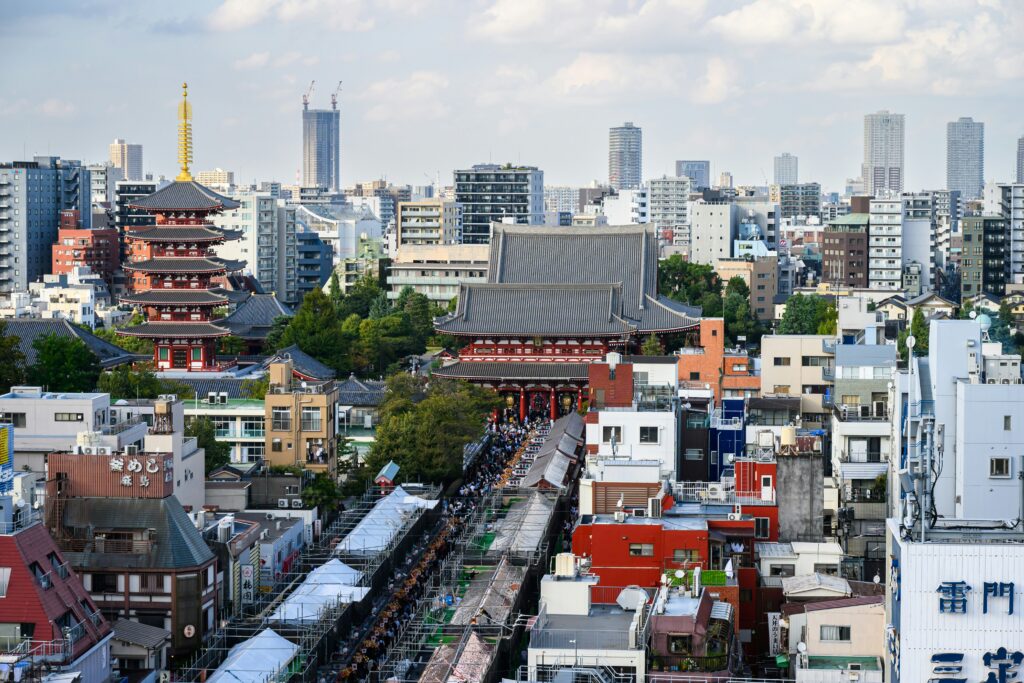 Aerial view of a cityscape featuring a mix of traditional and modern architecture. In the foreground, there are colorful low-rise buildings and a busy street lined with market stalls covered by white canopies. In the middle, a large traditional temple with a prominent pagoda is surrounded by trees. In the background, modern high-rise buildings and skyscrapers are visible under a partly cloudy sky.