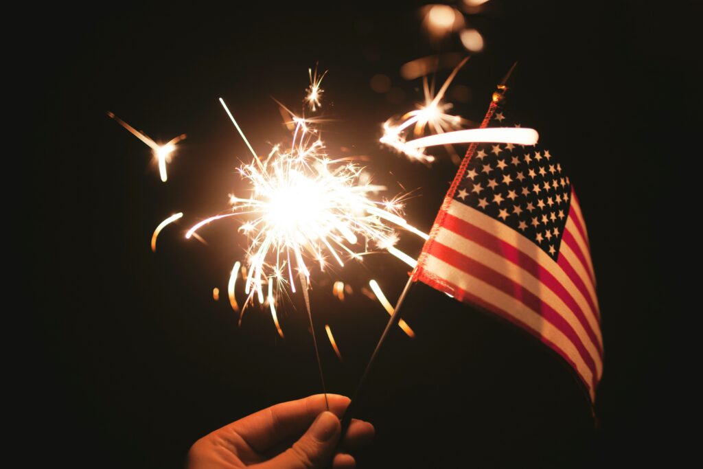A hand holding a lit sparkler and a small American flag against a dark background. The sparkler is emitting bright, glowing sparks.