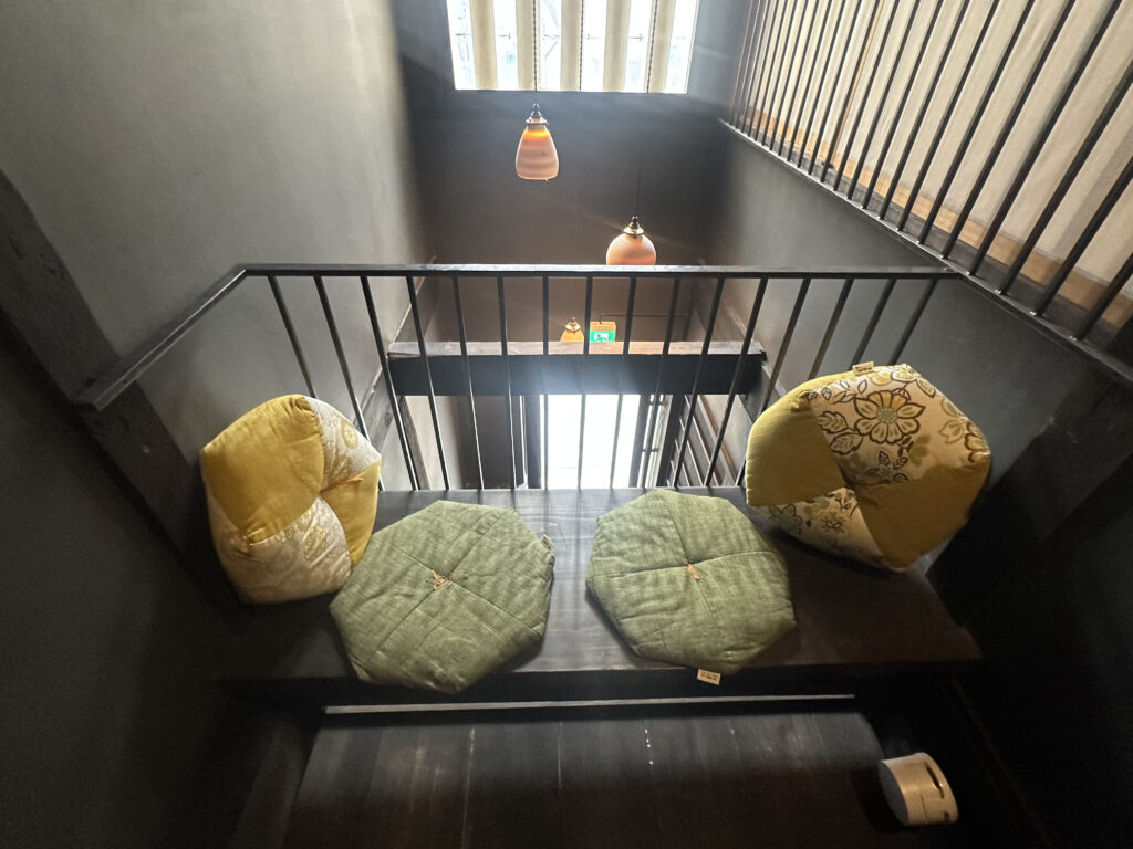 The image shows a small seating area on a landing with a dark wooden bench. There are three cushions on the bench: two are round with a patchwork design in yellow and floral patterns, and one is a green hexagonal cushion. The area is enclosed by metal railings, and there are two pendant lights hanging from the ceiling above. A small white device is on the floor near the bench.