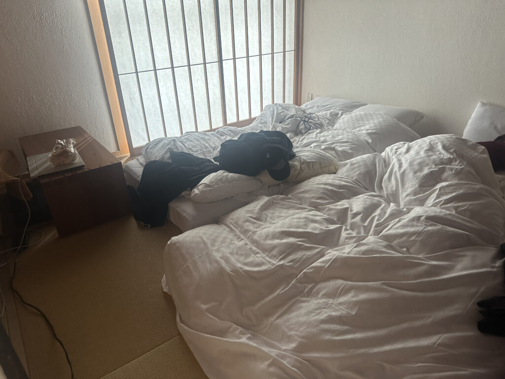 A room with a tatami mat floor features a low bed with white rumpled bedding. Clothes and a pair of shoes are scattered on the bed. A wooden side table holds a few items, including a wrapped package. A shoji screen window allows diffused light into the room.