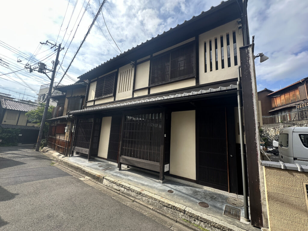 The image shows a traditional Japanese-style building on a quiet street. The building has a wooden facade with vertical slats and a tiled roof. There are power lines and a utility pole visible on the left side of the image. The sky is partly cloudy, and there is a small vehicle parked on the right side.