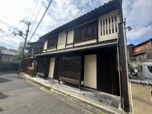 The image shows a traditional Japanese-style building on a quiet street. The building has a wooden facade with vertical slats and a tiled roof. There are power lines and a utility pole visible on the left side of the image. The sky is partly cloudy, and there is a small vehicle parked on the right side.