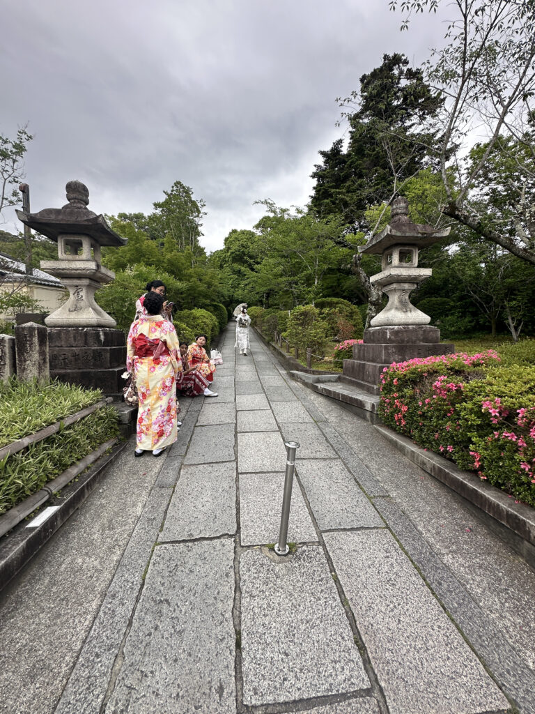 A stone pathway lined with greenery and traditional stone lanterns leads into the distance. Several people wearing colorful kimonos are walking along the path. The sky is overcast, and the surrounding area is lush with trees and bushes.
