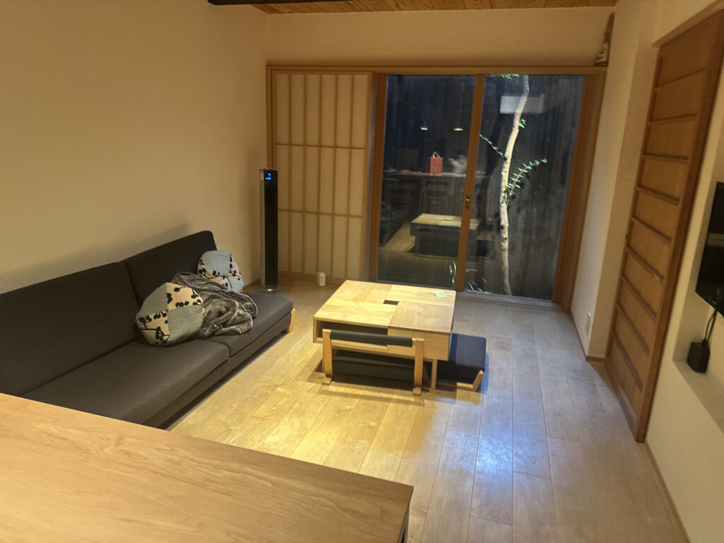 The image shows a cozy living room with a minimalist design. There is a dark gray sofa on the left with patterned cushions and a blanket. A wooden coffee table is in the center of the room, and a sliding glass door with a view of a small outdoor area is at the back. The floor is made of light wood, and there is a tall, slim fan or heater next to the sofa. The room has a warm, inviting atmosphere.
