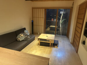 The image shows a cozy living room with a minimalist design. There is a dark gray sofa on the left with patterned cushions and a blanket. A wooden coffee table is in the center of the room, and a sliding glass door with a view of a small outdoor area is at the back. The floor is made of light wood, and there is a tall, slim fan or heater next to the sofa. The room has a warm, inviting atmosphere.