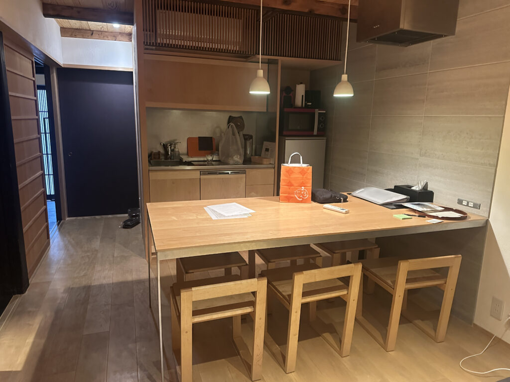 The image shows a modern kitchen with a wooden dining table and four matching chairs. The table has various items on it, including a bright orange shopping bag, papers, and a few other objects. The kitchen area in the background features a sink, a microwave, and some kitchen appliances. Two pendant lights hang above the table, and the room has wooden and neutral-toned walls and flooring.