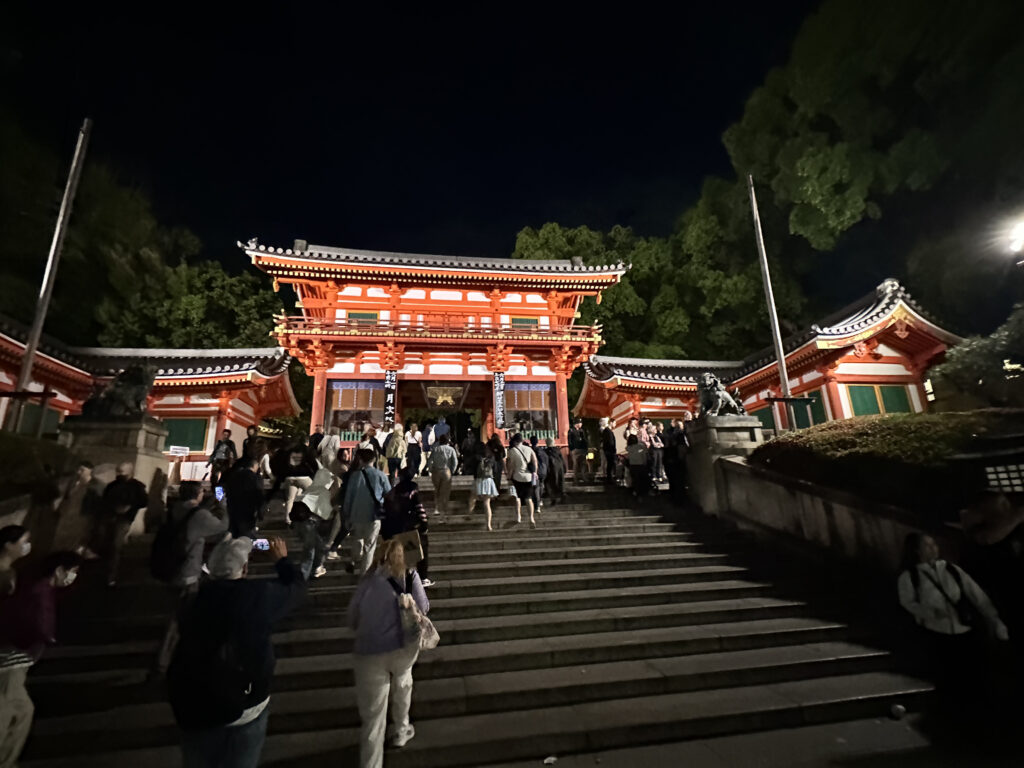 The image shows a large, illuminated traditional Japanese gate at night, with a crowd of people walking up the steps towards it. The gate is painted in red and white, with intricate architectural details. There are trees in the background, and the scene is well-lit, highlighting the structure and the people.