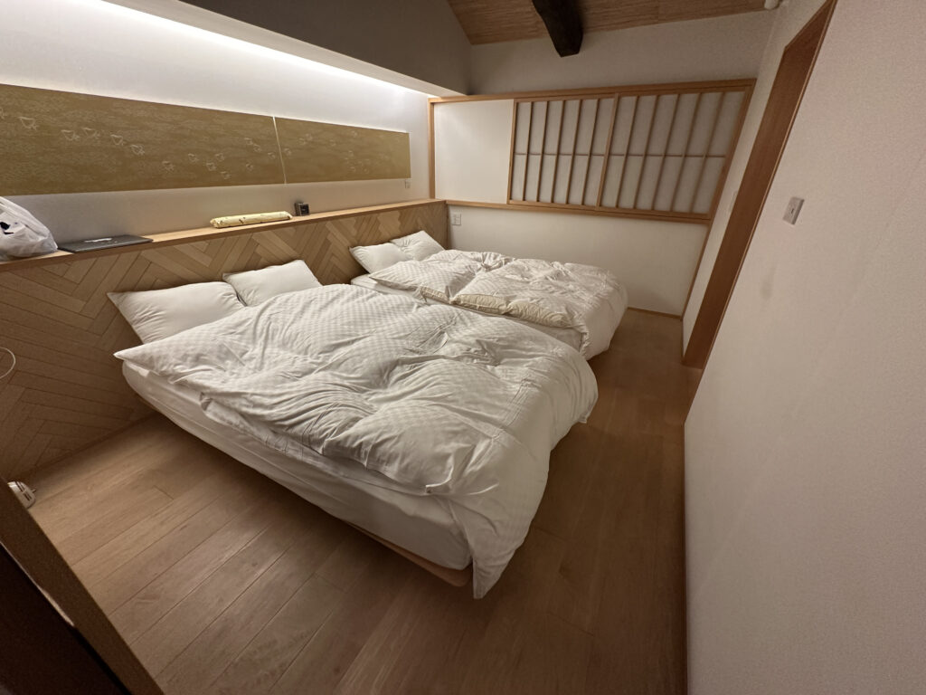 The image shows a minimalist bedroom with two low beds placed side by side on a wooden floor. The beds have white bedding and pillows. The room features a wooden accent wall with a shelf above the beds, holding a few items. A shoji-style sliding door is visible on the right side, adding a traditional Japanese touch to the room. The lighting is soft and warm, creating a cozy atmosphere.