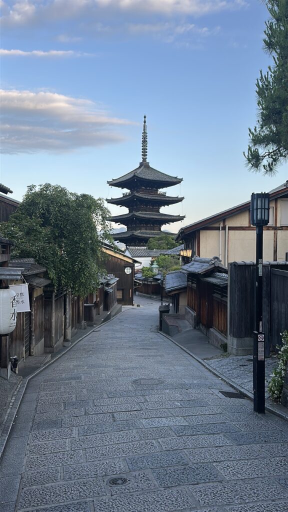 A narrow, cobblestone street lined with traditional wooden buildings leads to a tall, multi-tiered pagoda in the distance. The sky is clear with a few clouds, and there are trees and a lamppost along the street.