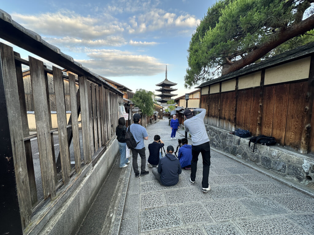 A group of people is gathered on a stone-paved street, taking photos of a person posing in the distance. The scene is set in a traditional area with wooden buildings on either side. In the background, a pagoda rises against a partly cloudy sky. A large tree extends over the right side of the path.