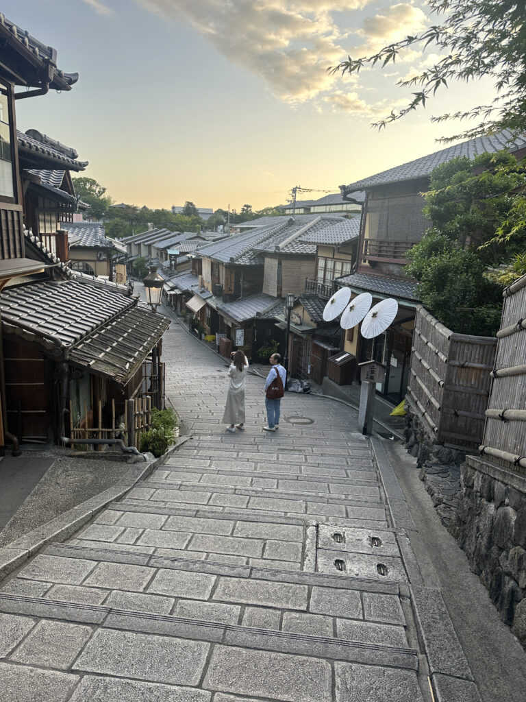 A narrow, stone-paved street in a traditional Japanese neighborhood, lined with wooden buildings featuring tiled roofs. Two people are walking down the street, and the sky is partly cloudy with a warm, golden hue from the setting sun. There are trees and greenery visible in the background.